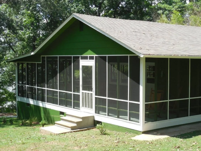 Green screened porch on a small house with steps and a sloped roof, surrounded by trees.