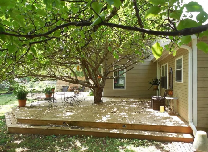 Wooden backyard deck under leafy trees beside a tan house, with patio table and chairs.