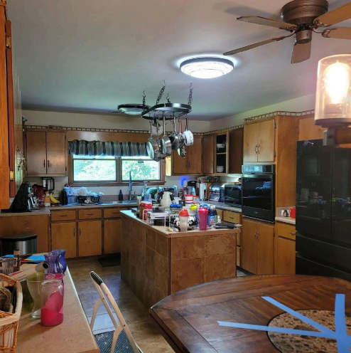 A cluttered kitchen with wood cabinets, a center island, hanging light, and a ceiling fan.