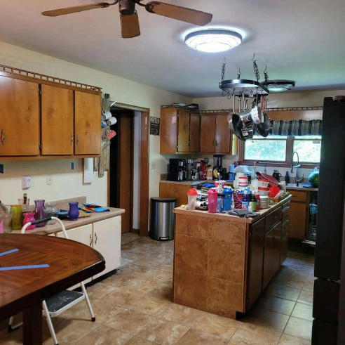 Cluttered kitchen with wooden cabinets, island, ceiling fan, and breakfast nook under a bright skylight.