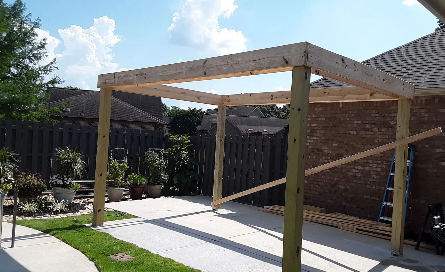 Backyard patio with a wooden pergola frame over a concrete slab beside a brick house and garden fence
