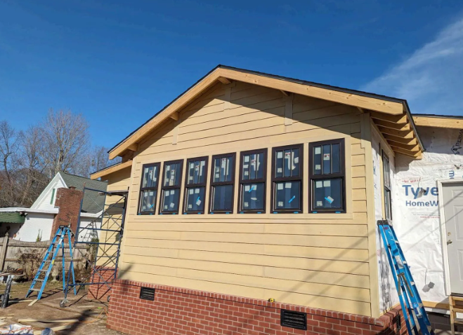 Tan house under construction with blue window wraps and a brick foundation under a clear sky