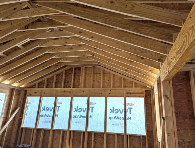 Unfinished wooden attic framing with exposed rafters and insulation-wrapped windows under construction