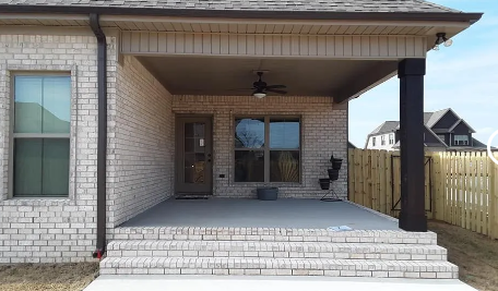 Front porch of a brick house with steps, columns, a ceiling fan, and a fenced yard.