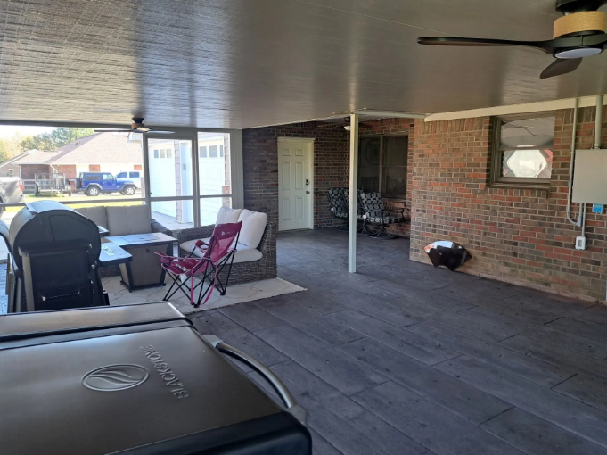 Covered patio with grill, table, and chairs beside a brick house and pool area