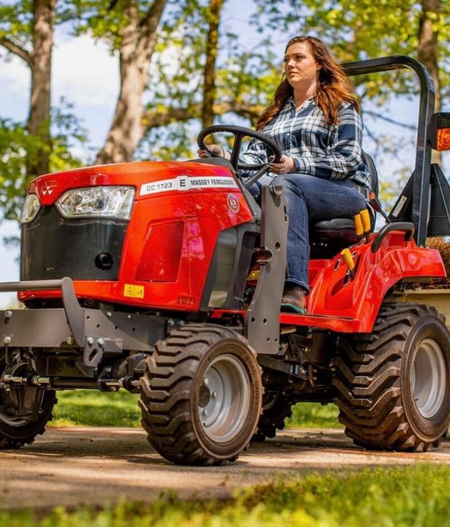 Woman driving a red tractor outdoors. She's wearing a plaid shirt and jeans. Bright, sunny day.
