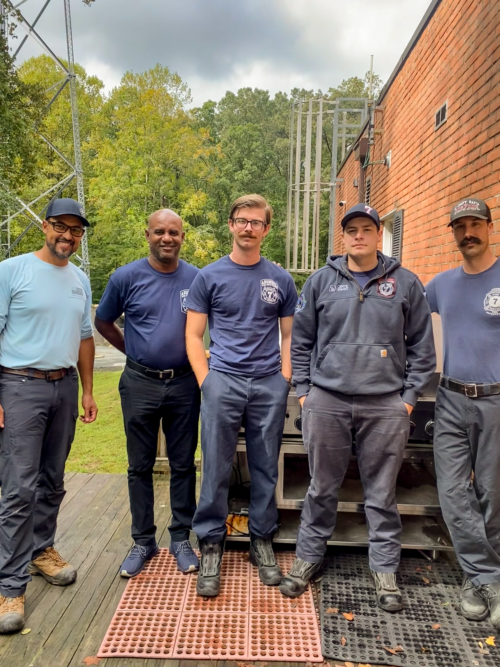 Five people in work attire stand outside. Brick building, metal structure, and greenery in the background.