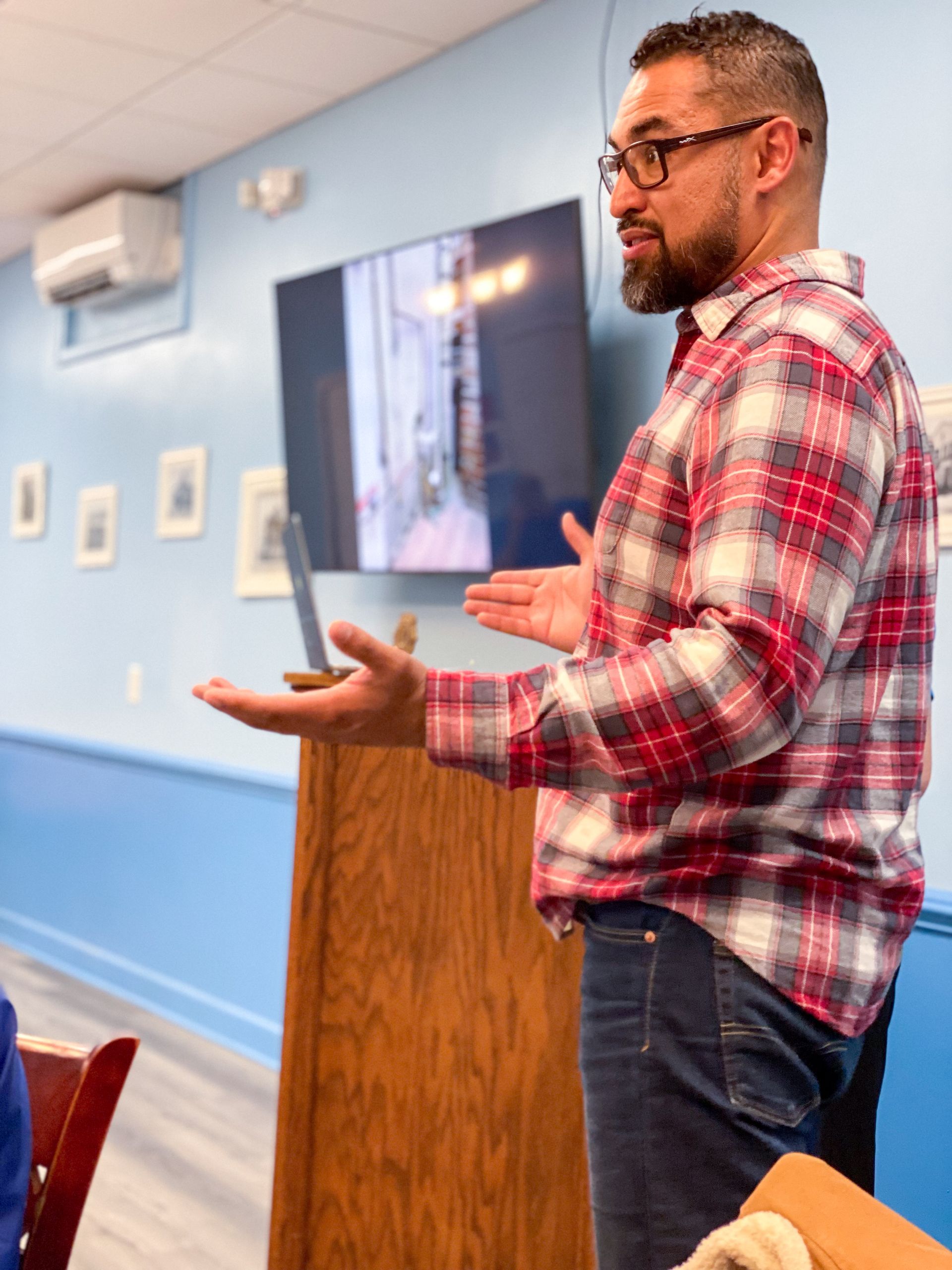 Man in plaid shirt speaking at a podium, gesturing toward a screen in a blue room.