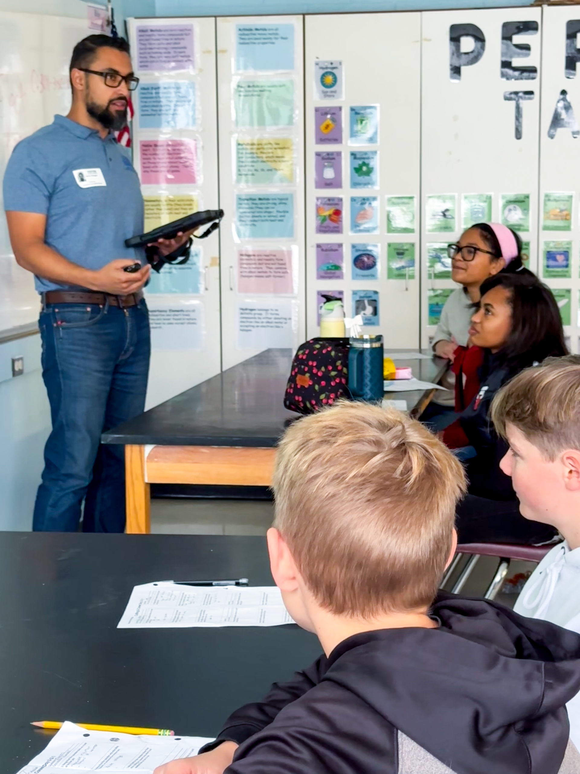 A teacher in a classroom, holding a tablet, speaking to students. 