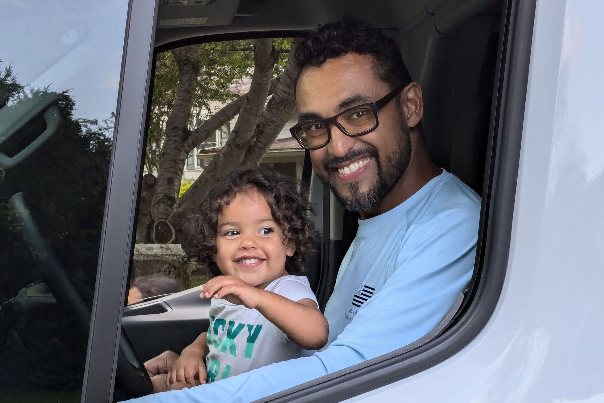Man and child smiling in a vehicle. Man in glasses, blue shirt, child in white top. Light setting.