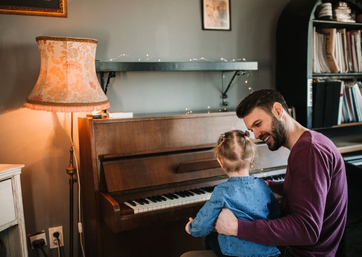Man and child seated at a piano in a cozy room with a lamp and shelves