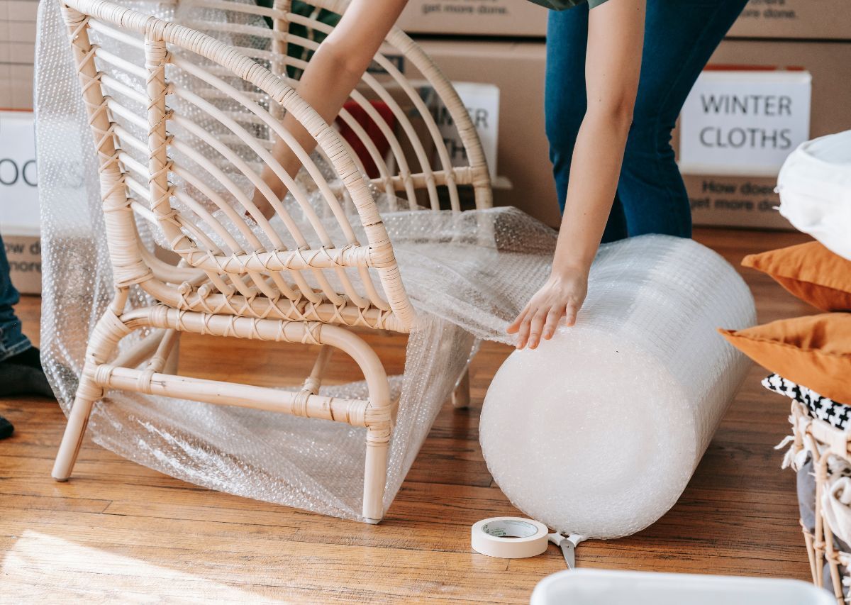 Person wrapping a white wicker chair in bubble wrap beside an orange couch during moving or storage