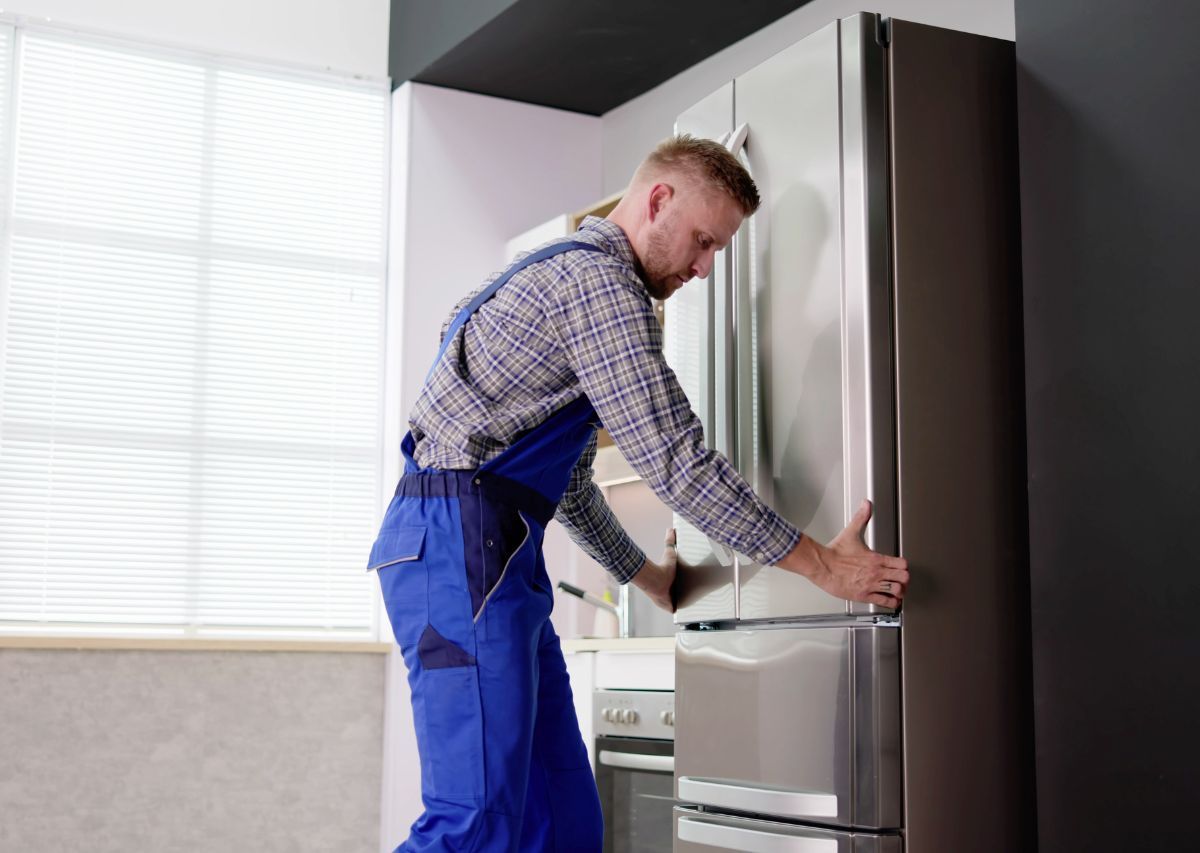 Technician in blue overalls adjusting a stainless steel refrigerator in a kitchen