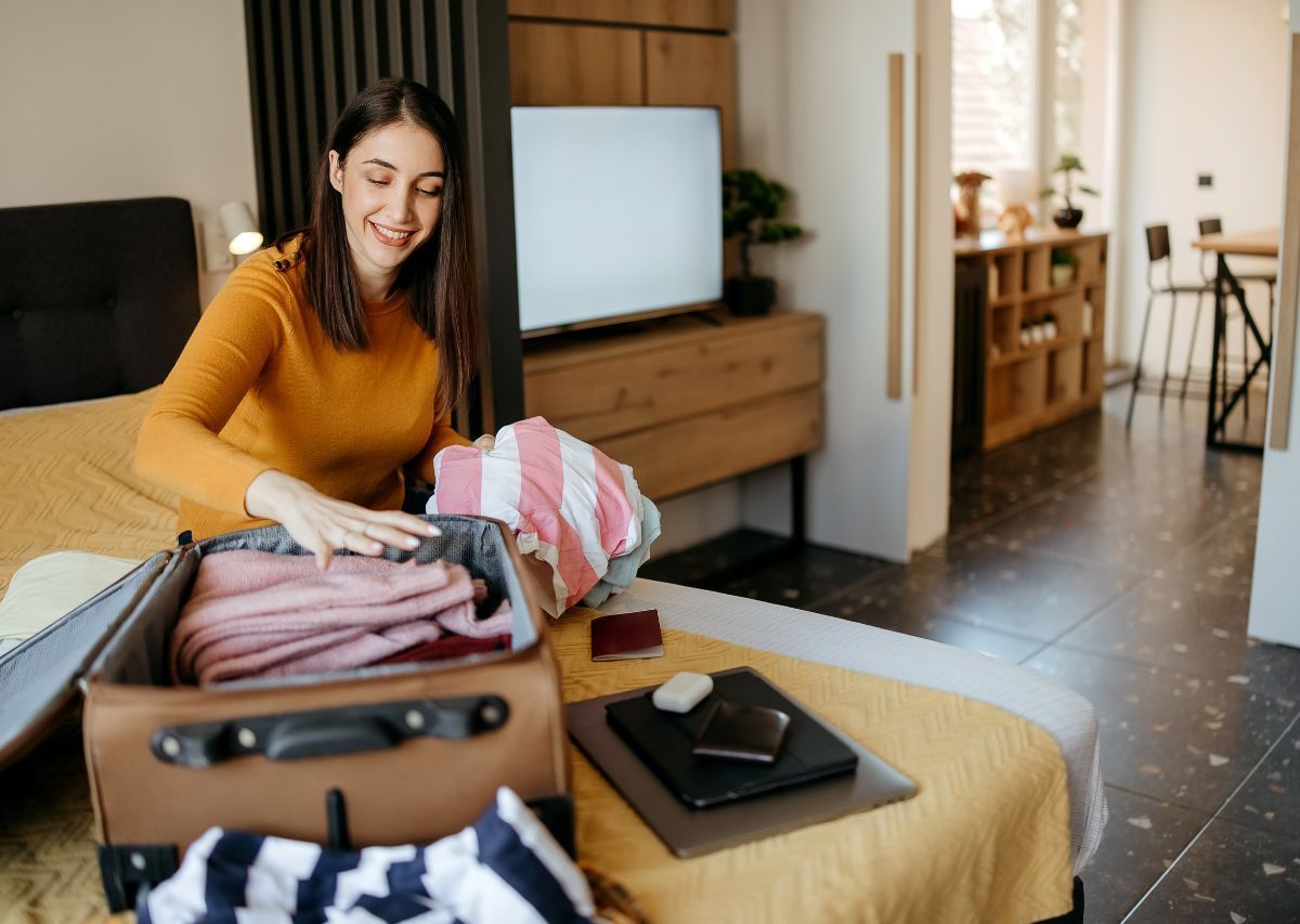 Woman packing clothes into an open suitcase on a bed in a bright bedroom