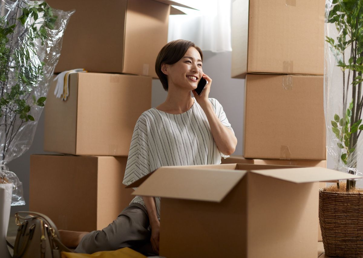 Woman on a phone call among stacked moving boxes in a sunlit room