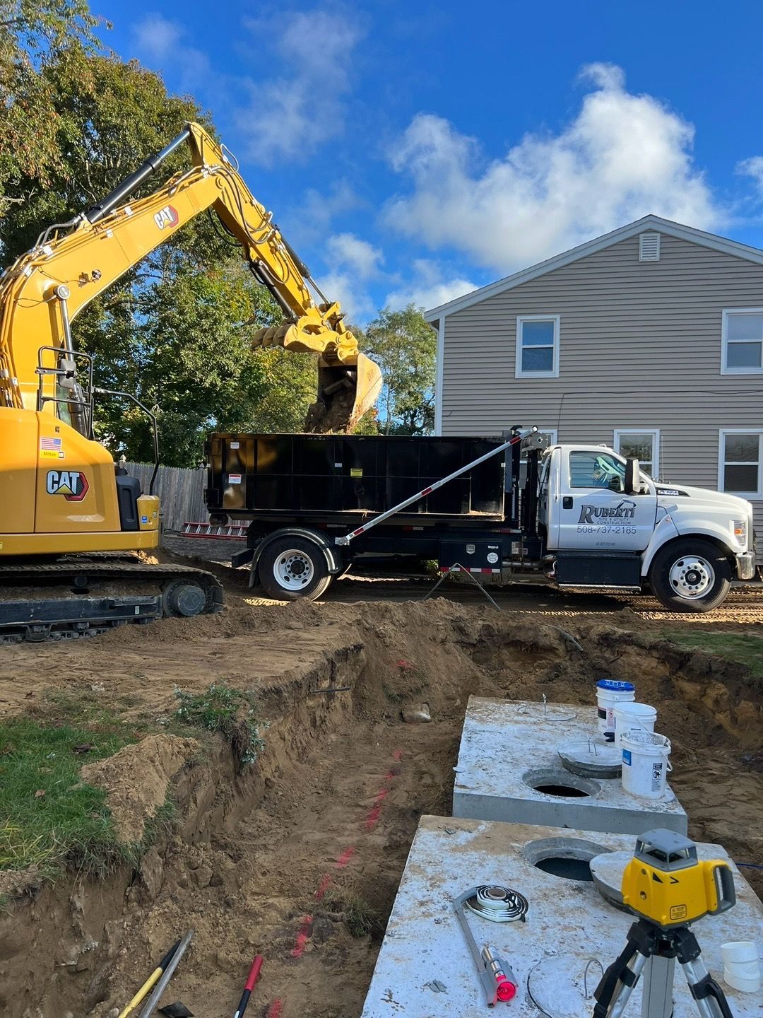 Yellow excavator loading dirt into a dump truck near a house, two concrete septic tanks in the foreground.
