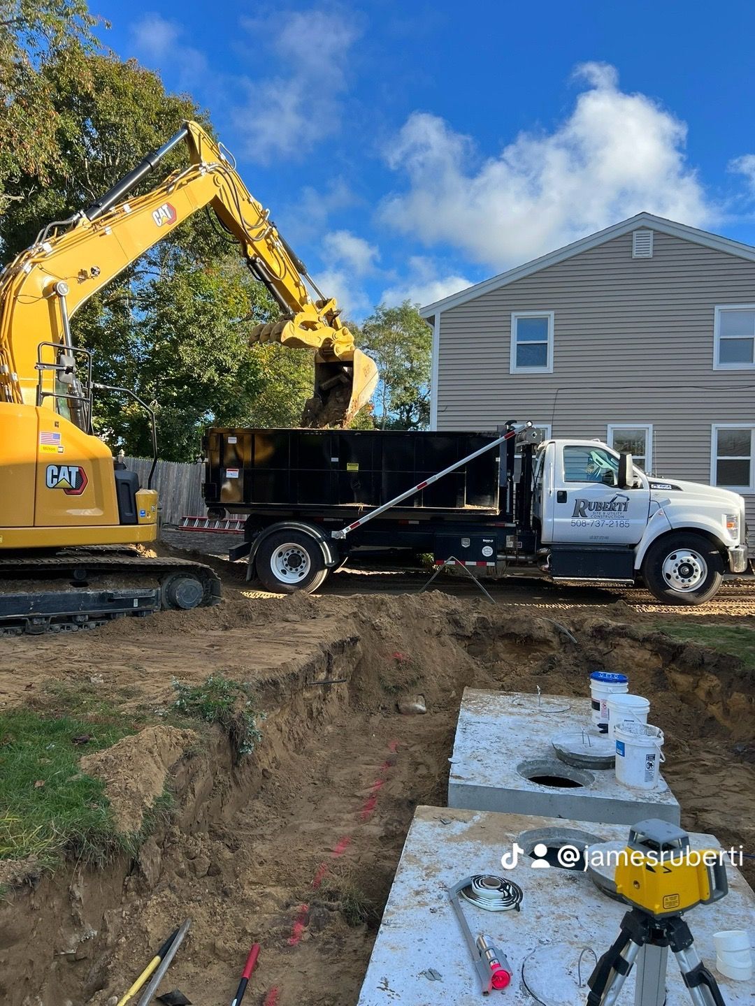Yellow excavator loading dirt into a black dump truck at a construction site near a house.