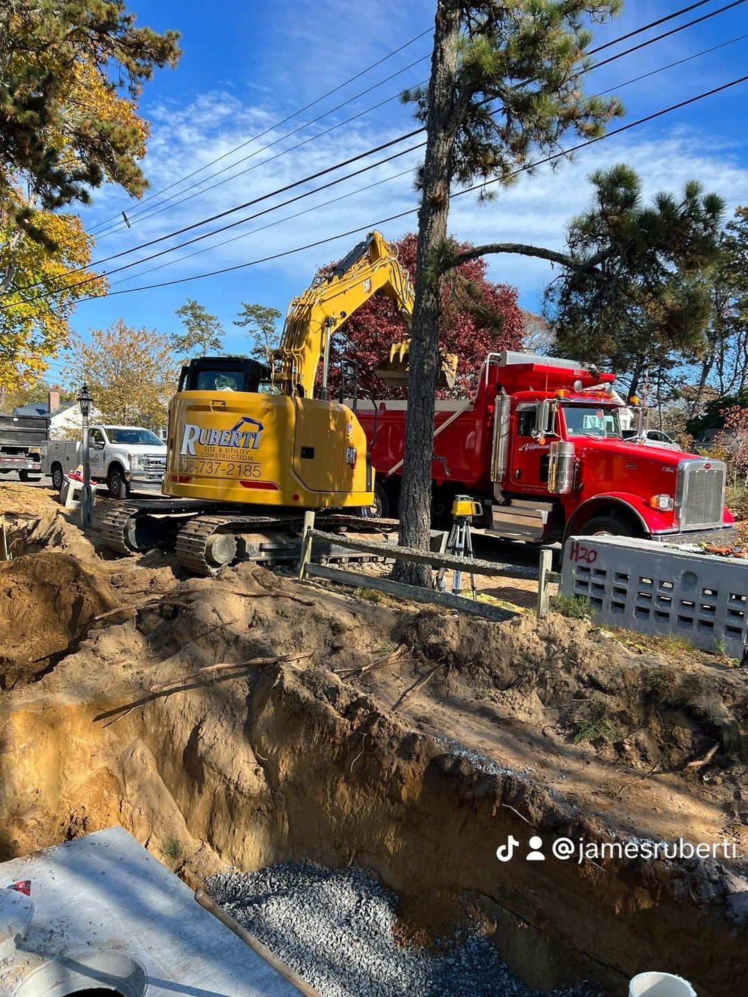 Yellow excavator working, red dump truck, digging near a road under a blue sky.
