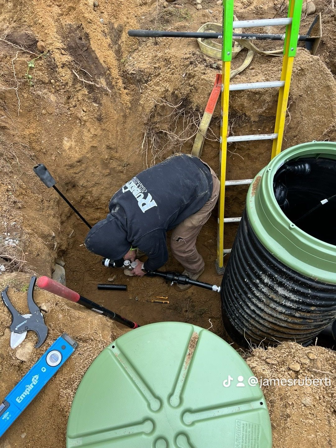 Man in a trench working on pipes, near a septic tank. Tools and a ladder are visible.