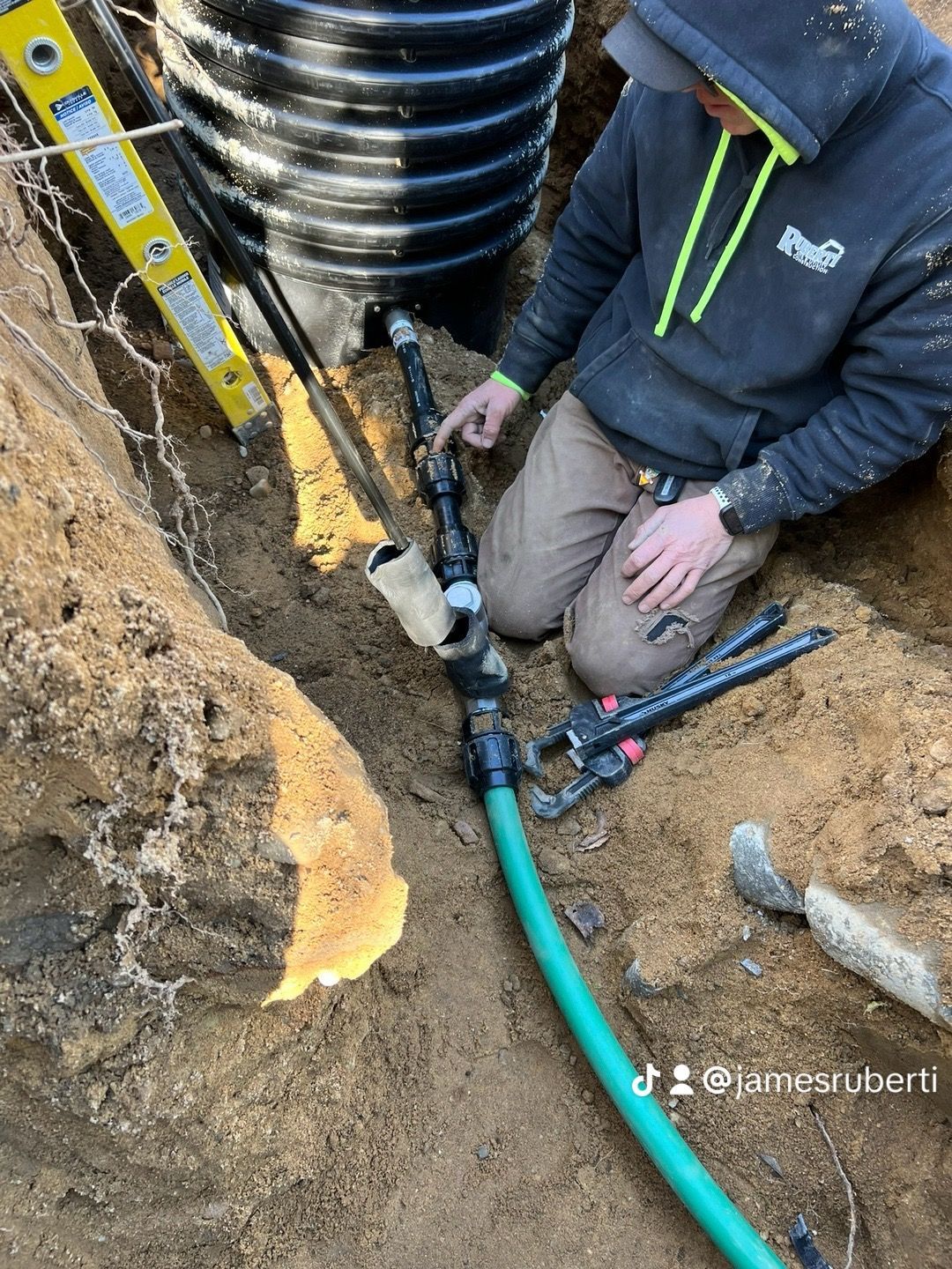 Person kneeling in a trench, pointing at black and white pipes connected to a black corrugated pipe. Green hose visible.