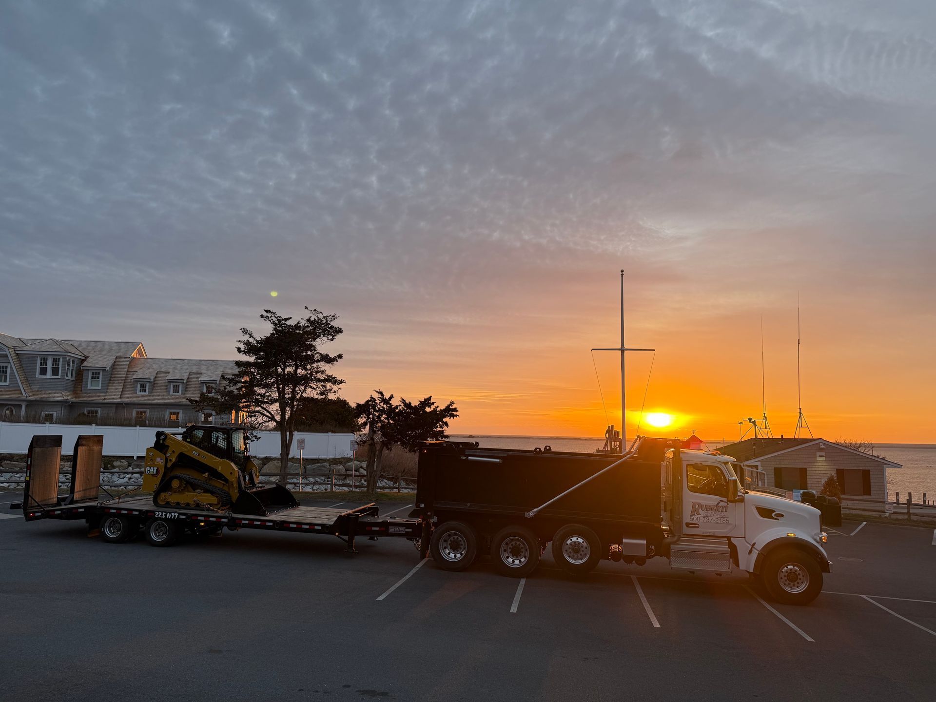 Dump truck with trailer carrying a yellow skid steer, parked at a waterfront as the sun sets.