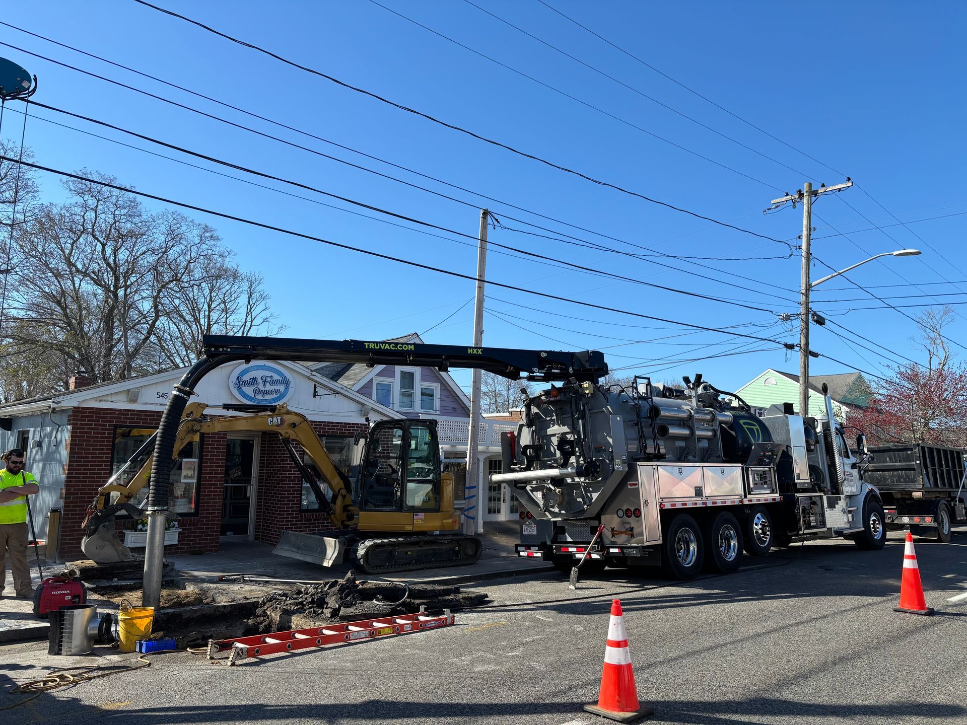 Construction site: excavator, vacuum truck, utility poles, street, house, workers. Blue sky.