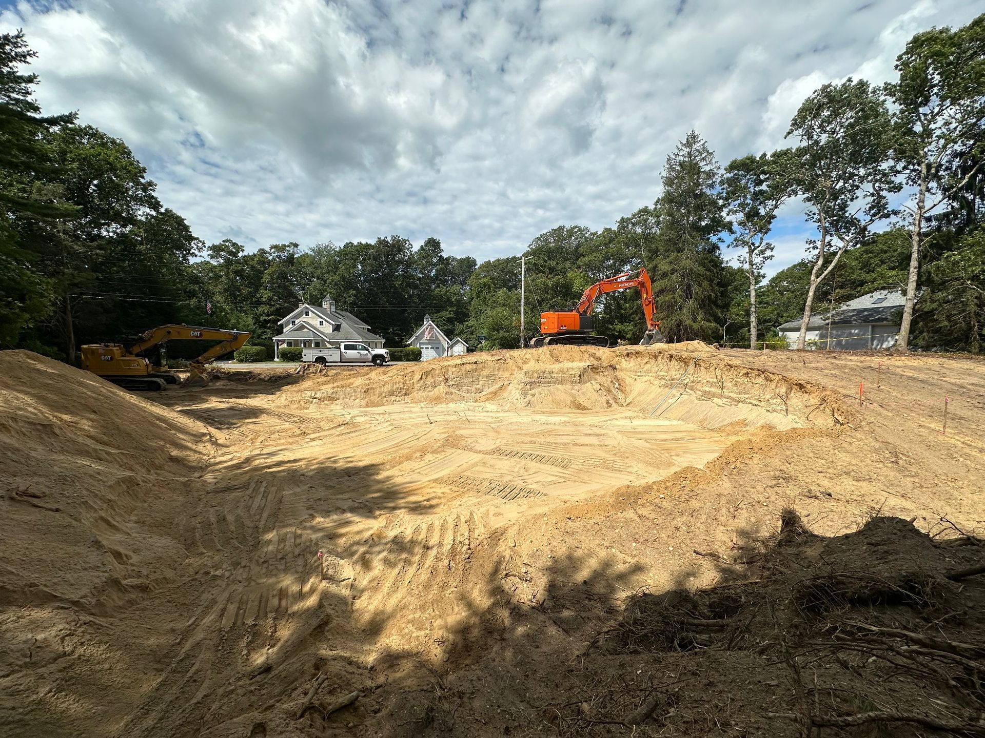 Construction site, excavated earth, two excavators, houses in background, cloudy sky.