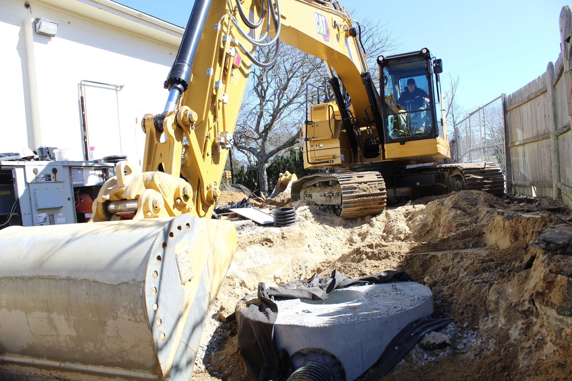 Yellow excavator digging near a building and fence, dirt and pipe visible.