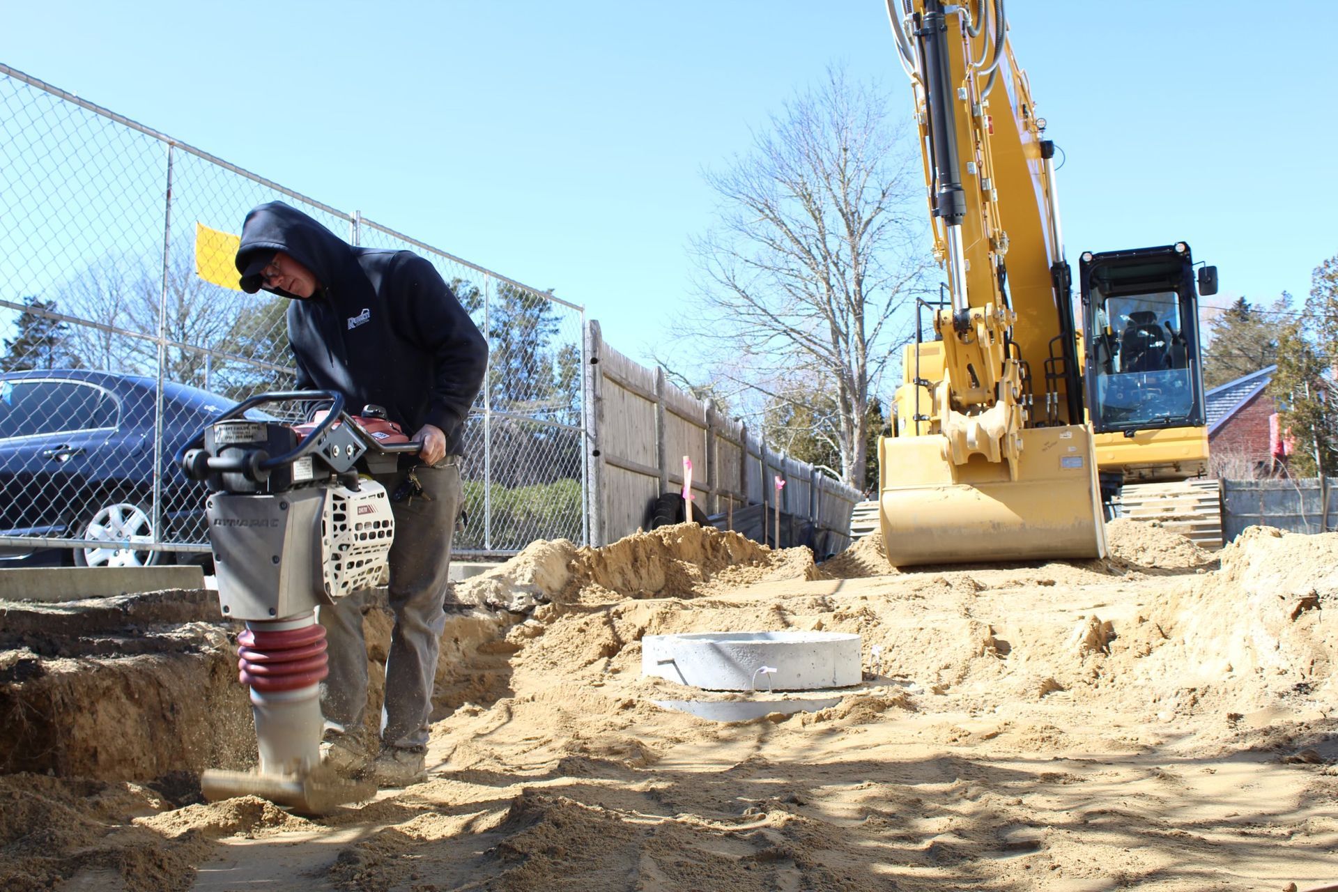 Person using a soil compactor in a construction site with an excavator.
