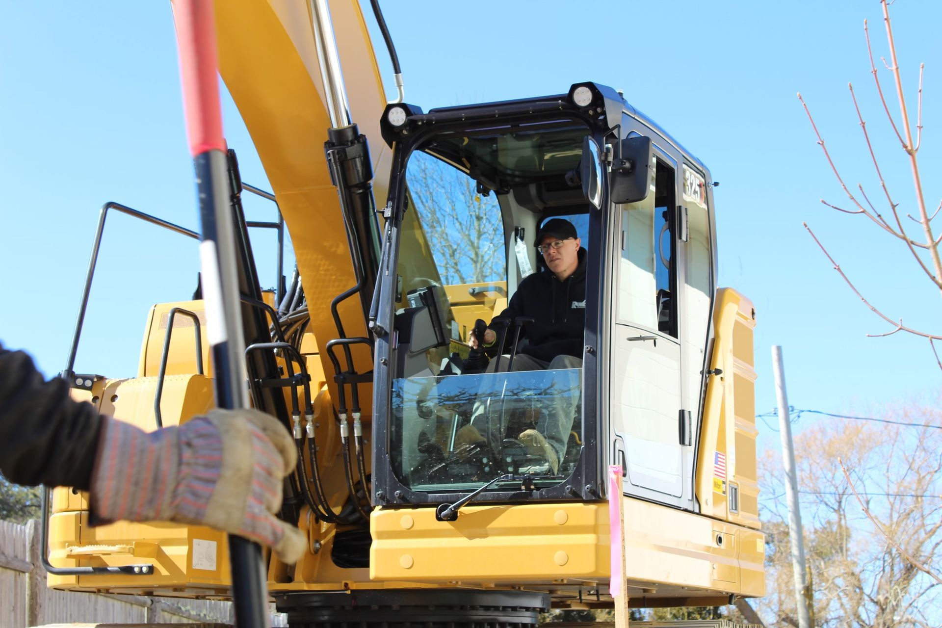 Person operating a yellow excavator. Someone holds a black tool, likely for guidance. Bright sunlight.