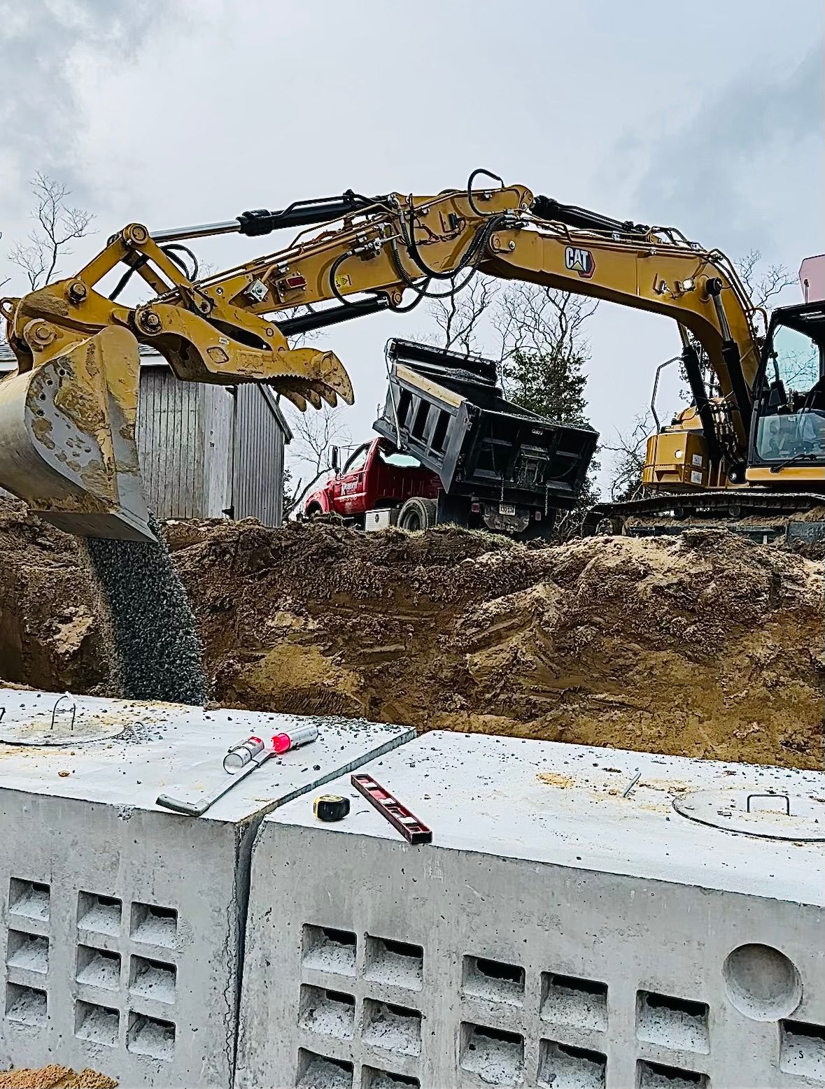 An excavator is dumping gravel into a concrete block structure in an earthen trench.