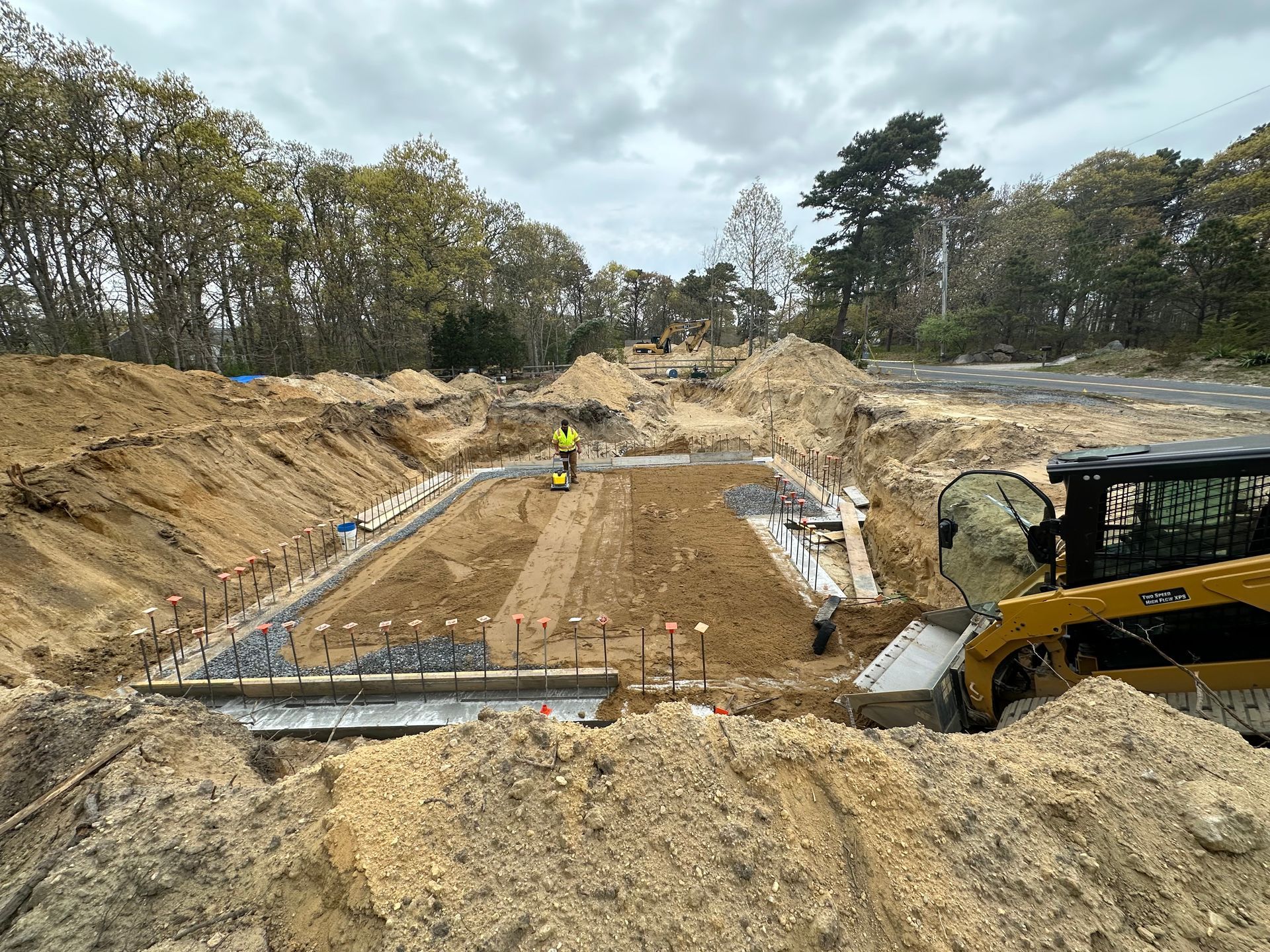 Construction site: Excavated area with concrete forms, a worker, and heavy machinery, under cloudy sky.