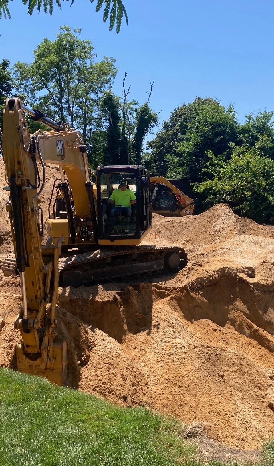 Yellow excavator digging in a dirt pit under a sunny sky; operator in cab.