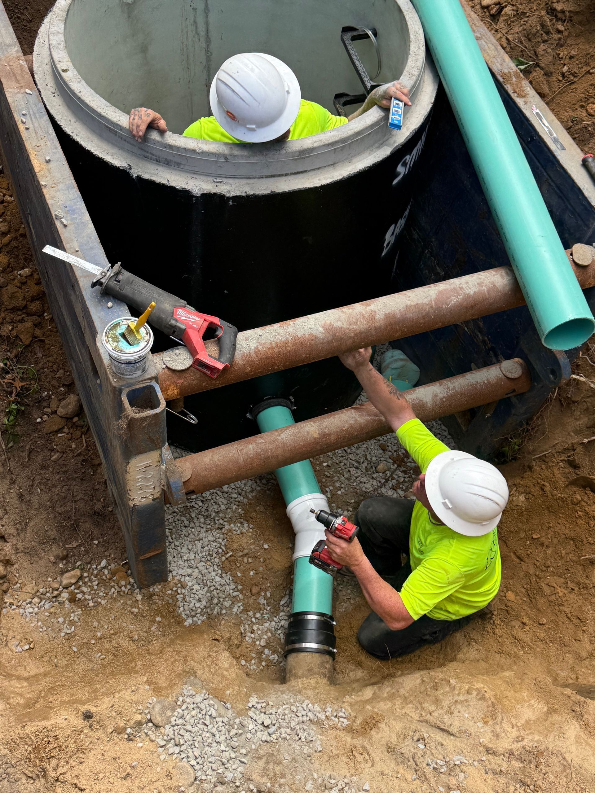 Two workers installing a pipe into a concrete manhole. They wear neon green shirts and hard hats, outdoors.