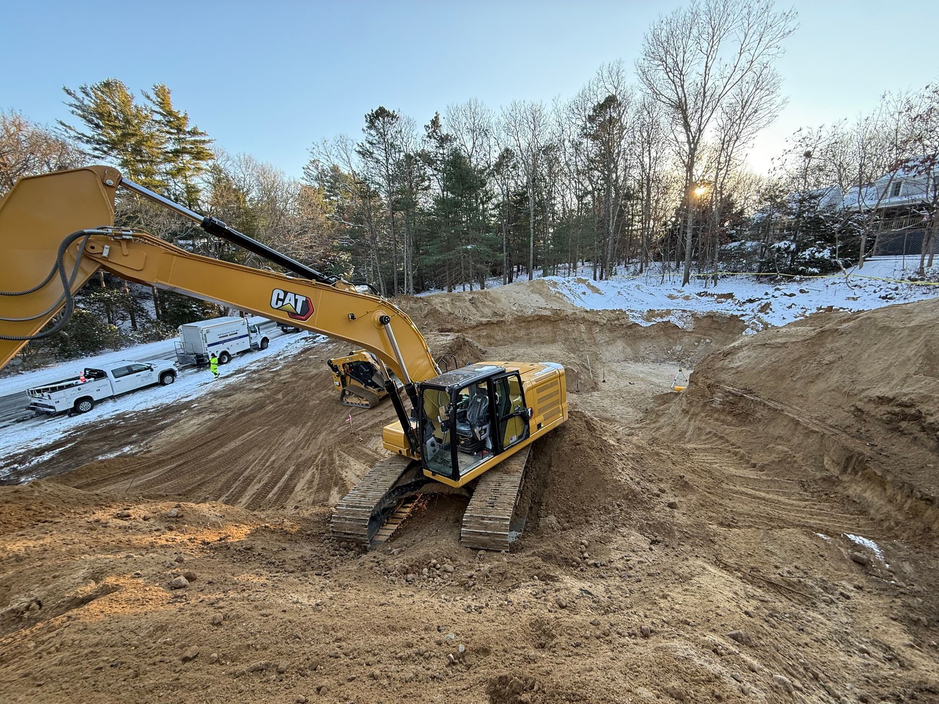 Yellow Caterpillar excavator digging in a dirt pit, with trees and a truck in the background. Snowy area.