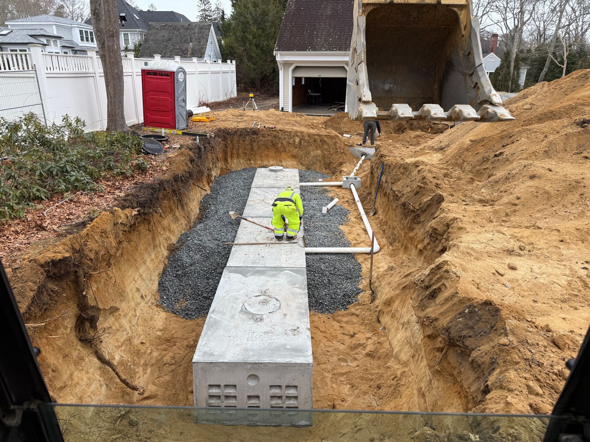 Construction site: Worker in yellow vest inspecting concrete septic system in trench, excavator in background.