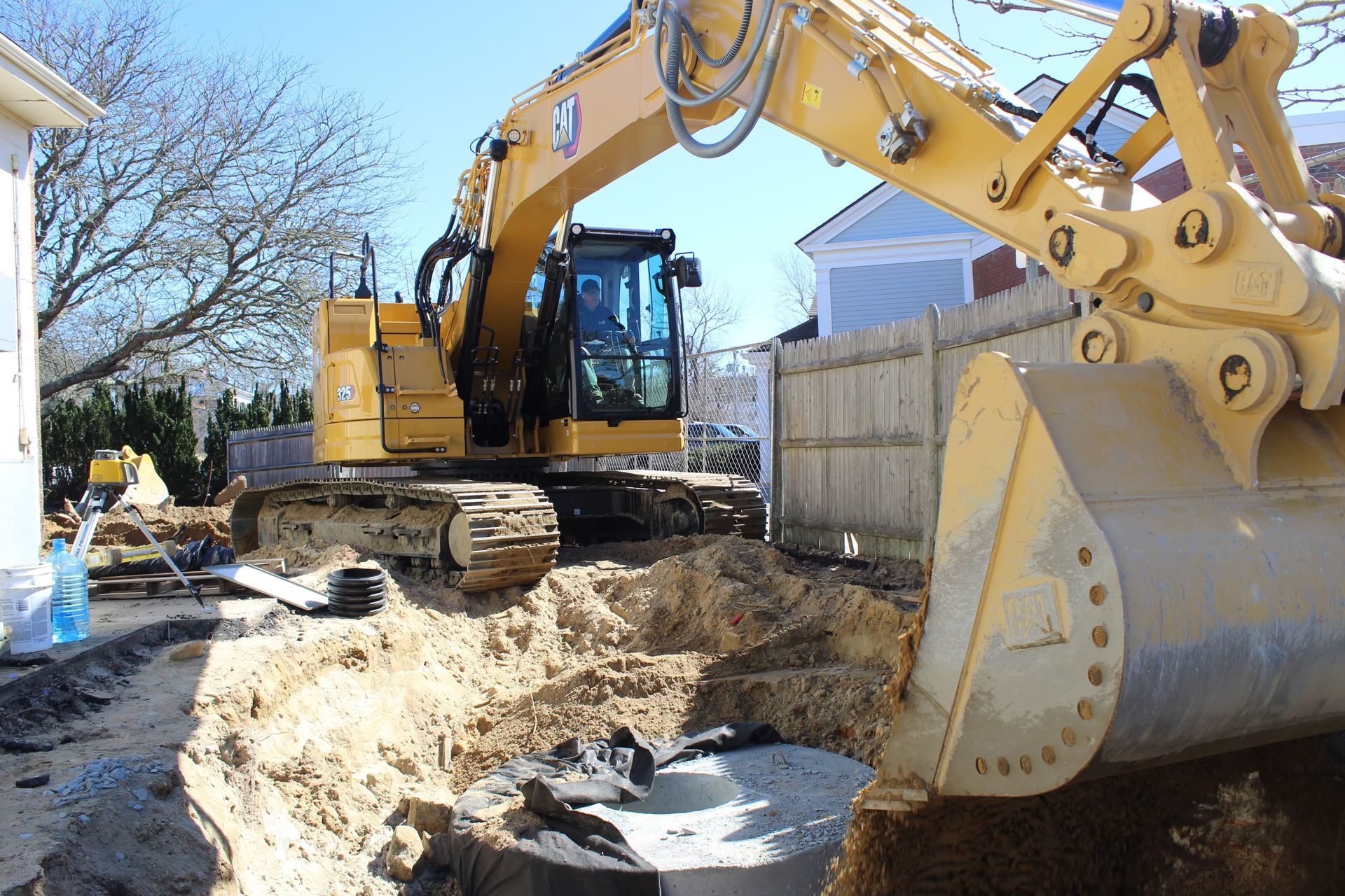 Yellow excavator digging near a wooden fence and building on a sunny day.
