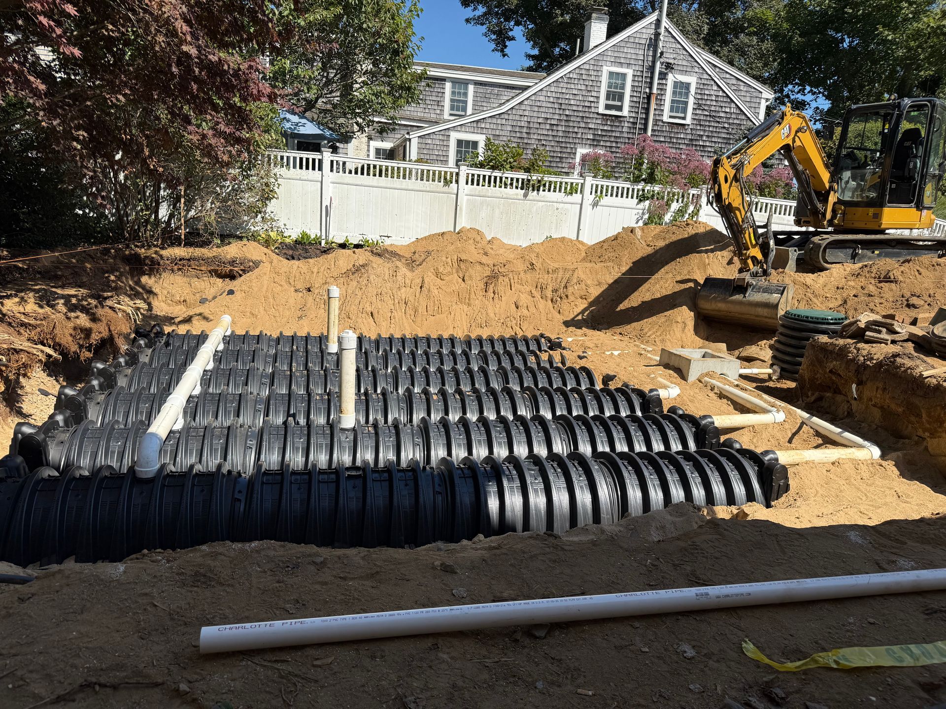 Excavated site with black septic tanks, white pipes, and a small excavator; house in background.