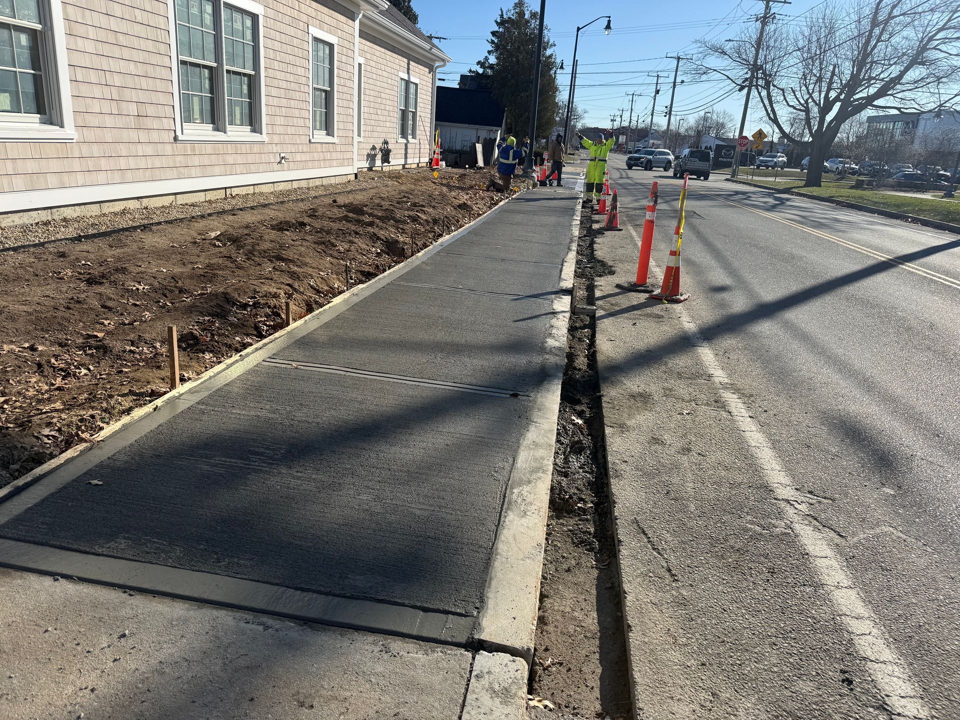 Sidewalk construction next to a road, orange cones, and a building on a sunny day.