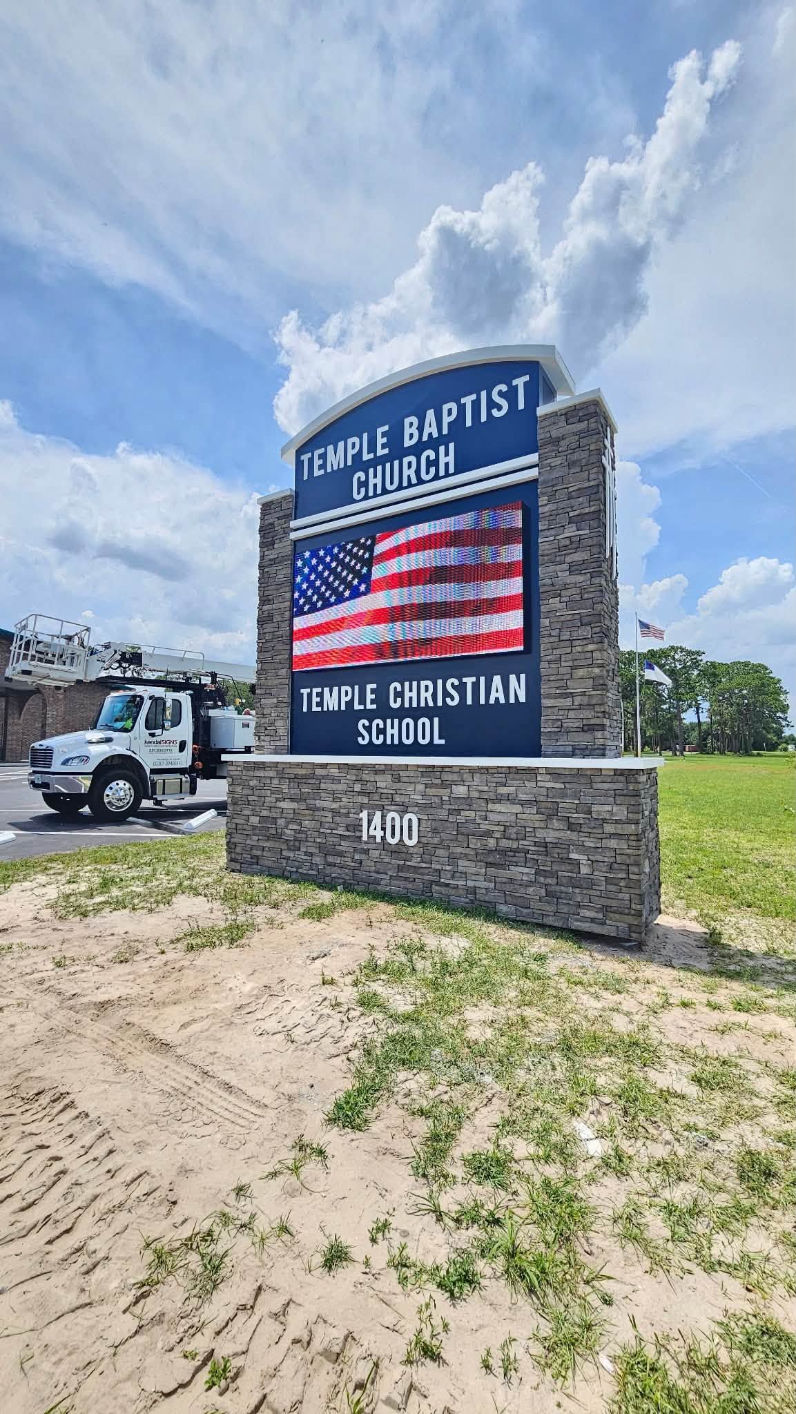Temple Baptist Church sign with American flag design on a sunny roadside field.