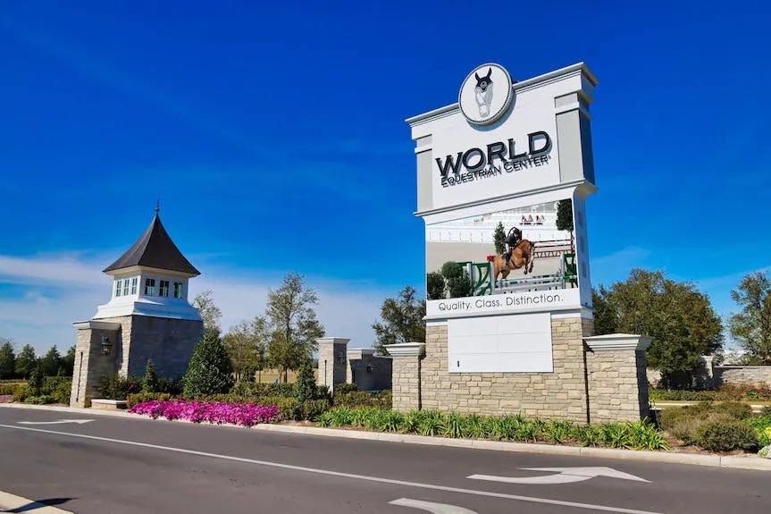 Stone entrance sign reading “WORLD” beside a roadside tower and pink flowers under a blue sky