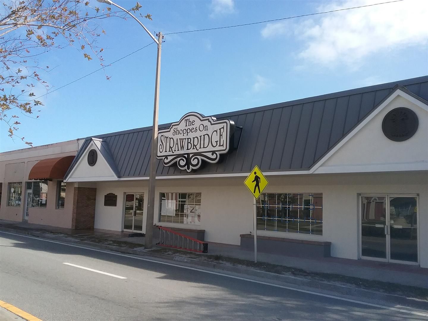 Street view of a white storefront with a black metal roof and sign, beside a crosswalk sign under a blue sky