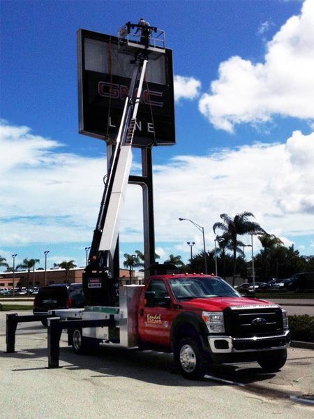 Red flatbed truck with a raised billboard sign beside a road under a blue sky