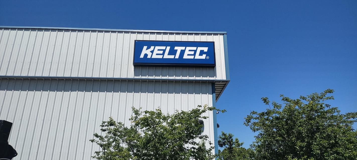 KELTEC sign on a light gray building against a clear blue sky and trees