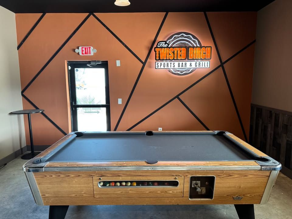 Empty pool hall with a pool table, orange walls, and a door with a neon exit sign.
