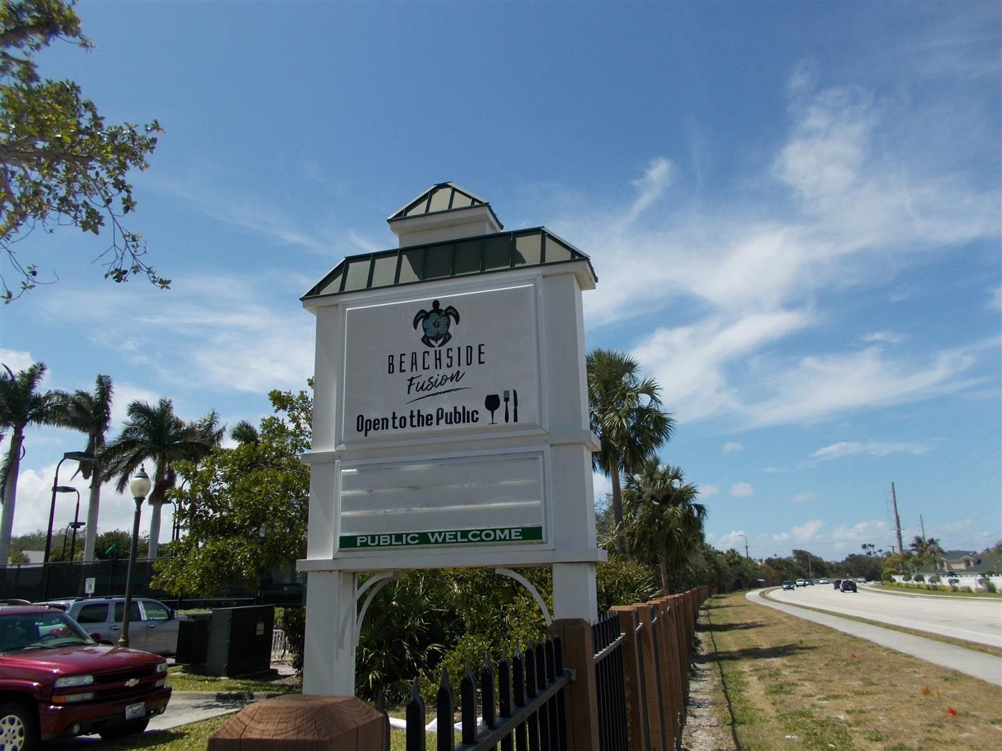 Entrance sign for The Villages at Palm Beach, beside a roadside with palm trees and blue sky
