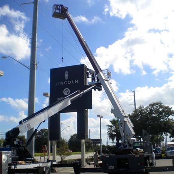 Two cranes lifting a large Lincoln sign at a dealership under a blue sky
