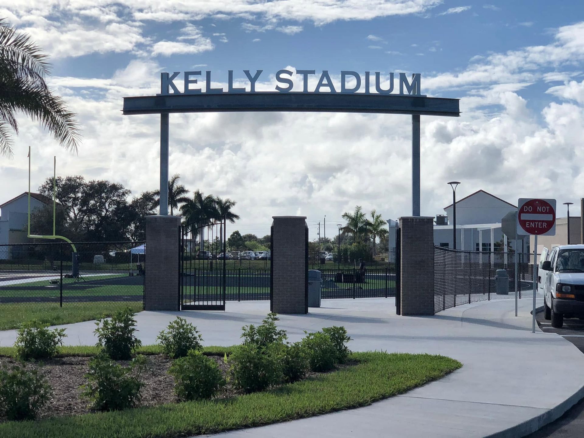 Kelly Stadium entrance gate with sign, palm trees, and a red stop sign on a sunny day