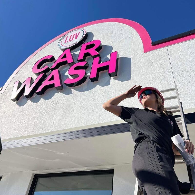 Woman standing outside a car wash sign, shading her eyes in bright sunlight