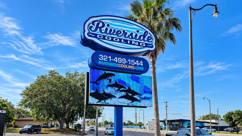 Riverside sign along a roadside under a bright blue sky, with cars and palm trees nearby